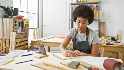 African american woman focusing on writing notes in a woodworking studio with tools and equipment...