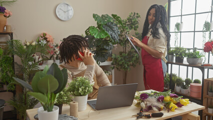 A woman florist arranges plants while a man sits at a laptop on a call in a flower shop interior.