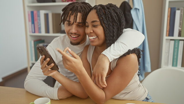 A smiling couple embraces while looking at a smartphone in a cozy living room setting, implying intimacy and digital connectivity.