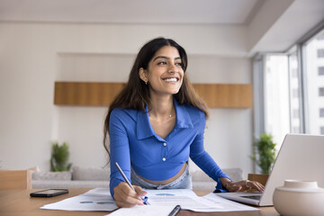 Cheerful young Indian project manager woman working on startup at home, bending over paper documents on table, writing notes, using laptop, looking away with toothy smile, thinking on creative ideas - 785307224