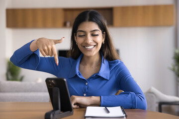 Happy attractive young Indian influencer woman shooting video review on mobile phone, sitting at work at home, speaking on conference call, showing finger at space downward at smartphone
