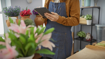 Hispanic man with beard uses tablet in flower shop surrounded by assorted plants, giving a modern entrepreneur vibe. © Krakenimages.com