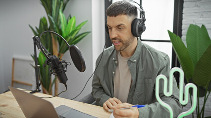 Handsome hispanic man with beard taking notes in a radio studio with microphone and headphones