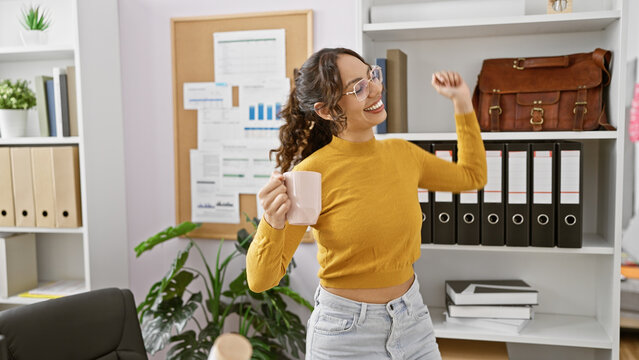 Joyful woman with glasses in a modern office celebrating a successful business achievement with a coffee mug.