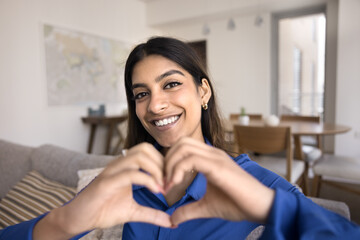 Cheerful young Indian influencer woman making finger heart gesture, expressing love, happiness, kindness, showing charity, friendship symbol to blog audience, smiling, looking at camera