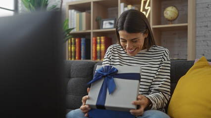 A cheerful hispanic woman enjoys unwrapping a gift in a cozy, book-filled living room.