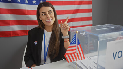 American young woman pointing indoors at electoral college with flag, ballot, vote sticker, and cheerful expression.