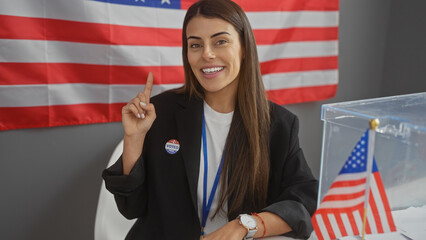 Smiling young hispanic woman with voter sticker pointing up in a room with american flags and...