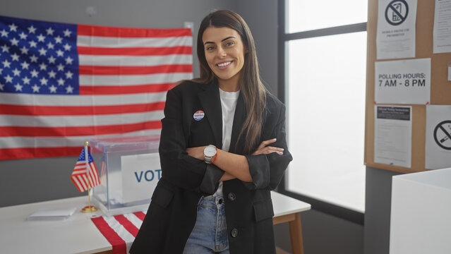A confident hispanic woman stands with crossed arms in an american electoral college room with a flag and voting signs. - Powered by Adobe