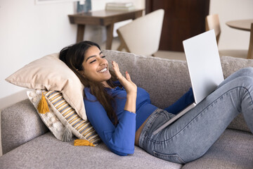Happy friendly young Indian woman waving hand hello at laptop, making hi greeting gesture, speaking on online video conference call, smiling, lying on comfortable home couch