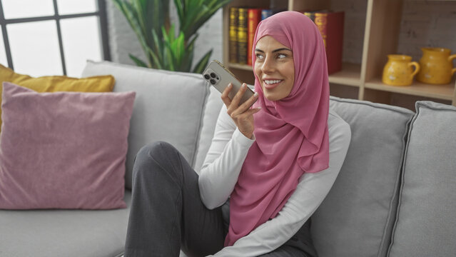 A young woman in a hijab sending a voice message on her phone at home, surrounded by cozy decor.