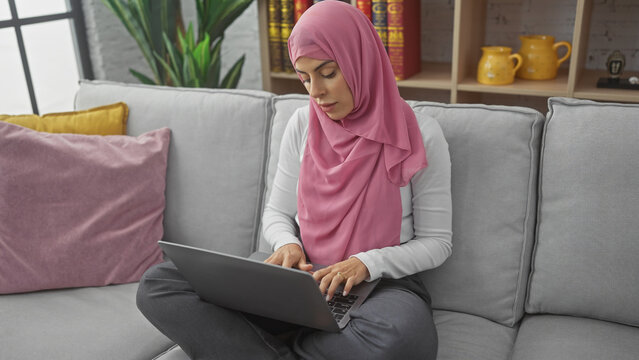A young woman in a hijab using a laptop on a couch indoors, surrounded by colorful pillows and home decor.