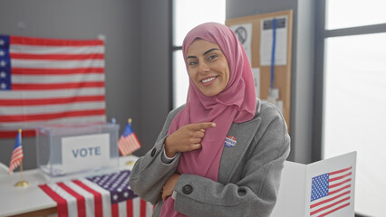 A confident young woman wearing a hijab points to her 'i voted' sticker in a us electoral college room adorned with american flags.