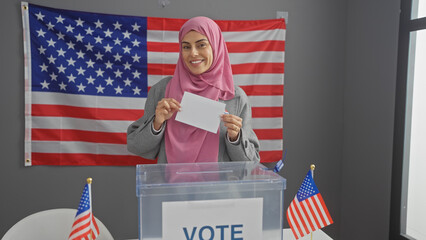 A smiling young hispanic woman wearing a hijab holds a ballot in a college electoral center with american flags. - obrazy, fototapety, plakaty