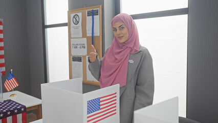 A young woman with a hijab gives a thumbs-up in a voting booth with american flags.