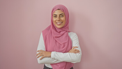 Confident young woman wearing hijab poses with arms crossed against a pink background, exuding...