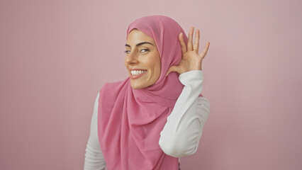 Smiling young hispanic woman in a pink hijab gestures listening isolated against a pink wall.