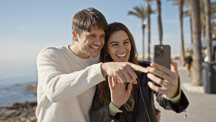 A smiling couple enjoys a sunny day at the seaside, taking a selfie with palm trees in the background.