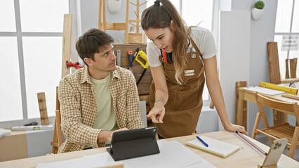 Man and woman discussing a project in a bright carpentry workshop with tools and wooden furniture.