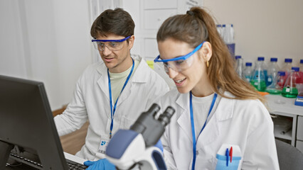 A man and woman in lab coats analyze data on a computer in a bright laboratory setting, indicative...