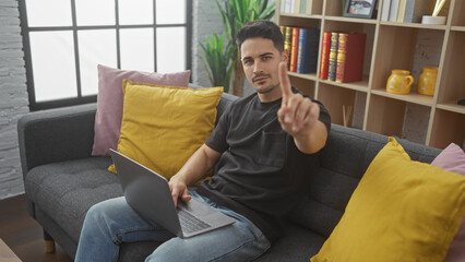 Handsome hispanic man with a beard gesturing no while sitting on a couch with a laptop in a modern...