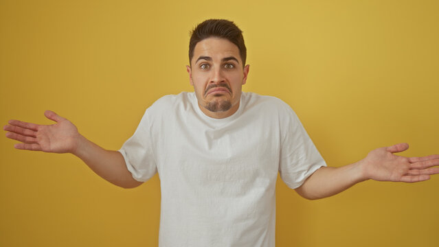 Hispanic man with beard in confusion against a yellow background, exhibiting a shrug in casual white clothing.