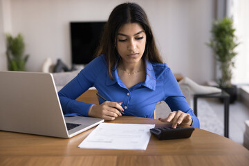 Serious young Indian accountant woman calculating domestic expenses, typing on calculator at laptop, paying bills, checking paper invoices. Freelancer working at home, counting budget - 785310814