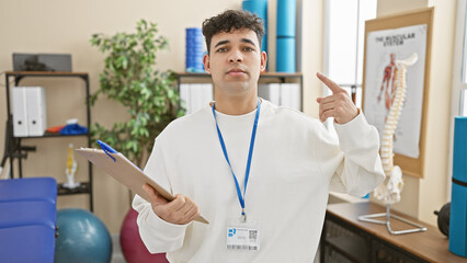A young man with a beard, wearing a badge, gesturing to his head in a physiotherapy clinic.