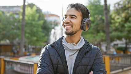 Smiling hispanic man with headphones enjoying music in a leafy city park.