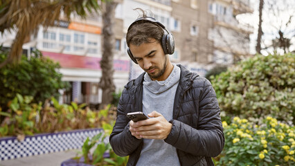 A young hispanic man with a beard, wearing headphones, looks at his phone in a lush city park.