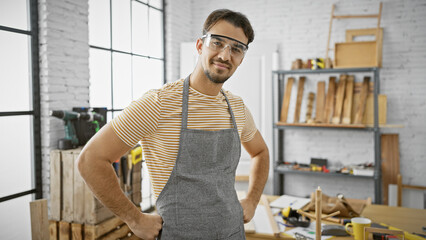 A confident young hispanic man with a beard wearing safety glasses and an apron poses in a...