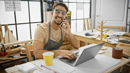 A smiling hispanic man with a beard in a woodworking workshop talks on the phone while using a...