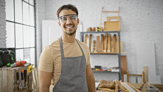 A smiling hispanic man with a beard wearing protective glasses and apron stands confidently in an organized carpentry workshop.