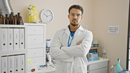 Confident hispanic man in lab coat standing in a medical laboratory with arms crossed.