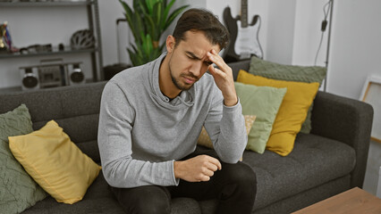 A young hispanic man with a beard appears stressed while sitting on a gray couch indoors, showcasing a modern home interior.