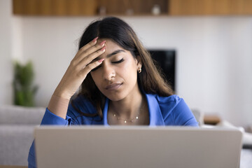 Exhausted overworked young Indian freelancer woman touching head with closed eyes at laptop,...