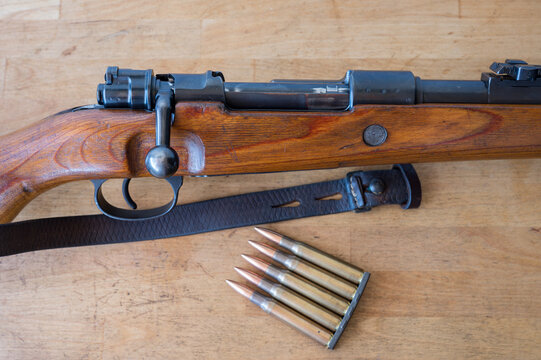 Detail of an ammunition clip with 5 30-06 caliber rifle bullets next to a Mauser-type bolt-action military rifle on a wooden table