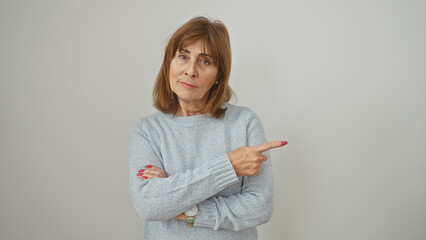 A sceptical middle-aged woman in a blue sweater points to the side against a white wall.