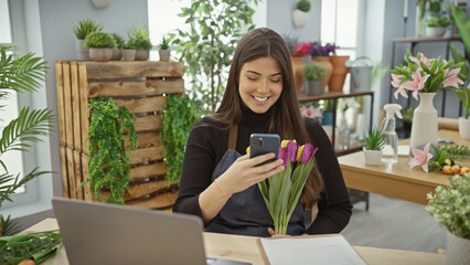 Smiling woman with flowers using smartphone in a vibrant indoor flower shop.