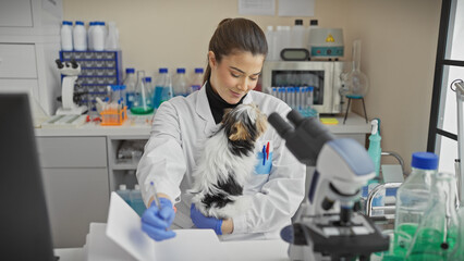 A young hispanic woman examines a biewer yorkshire terrier in a laboratory indoor setting.