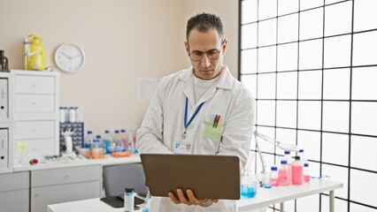 A focused hispanic man in a laboratory setting examines data on his laptop amidst medical equipment and glassware.