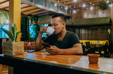 Relaxed asian man drinking coffee and checking social media on smart phone in the coffee shop while evening.