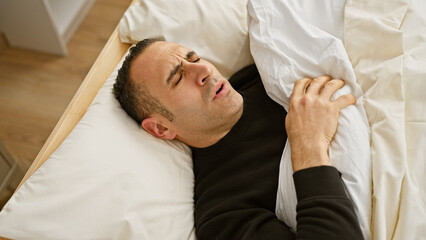 A distressed man grimacing in pain while lying in bed, portraying sickness or discomfort in a home bedroom setting.