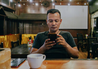 Asian Indonesian man sitting relaxed alone in coffee shop or bar at night while reading the news on his cellphone.