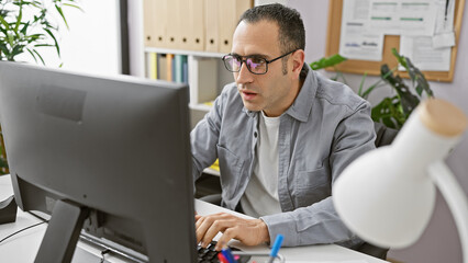 Hispanic man working concentrated at his desk in a well-lit modern office, using a computer and wearing casual attire