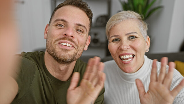 A cheerful man and woman smiling and waving at the camera in a well-lit modern living room interior