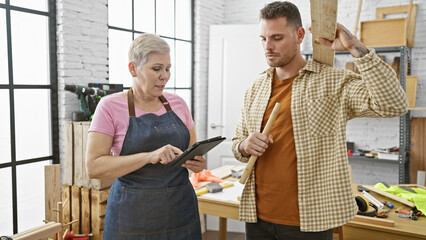 A woman and man review plans on a tablet in a well-lit carpentry workshop, surrounded by tools and wood.