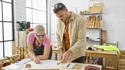 A man and woman collaboratively work in a light-filled carpentry workshop surrounded by tools and...