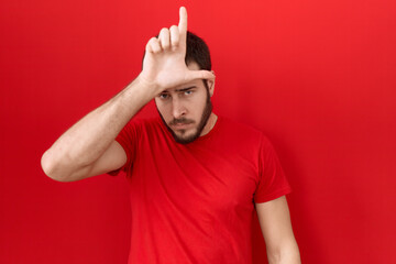 Young hispanic man wearing casual red t shirt making fun of people with fingers on forehead doing...