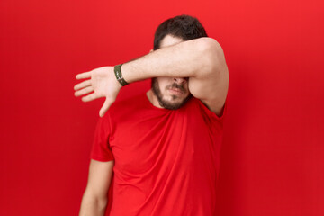 Young hispanic man wearing casual red t shirt covering eyes with arm, looking serious and sad....